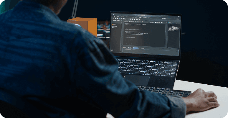 A person sits at a desk using a laptop that displays a dark-themed code editor.