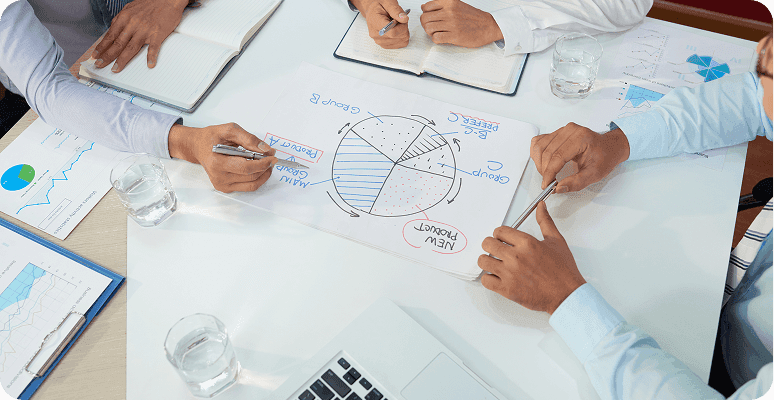Several people sit around a table collaborating over documents, charts, and a laptop.