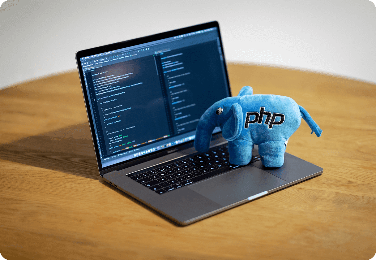 A laptop displaying code with a blue plush PHP elephant resting on the keyboard
