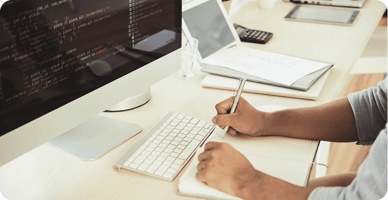 Person taking notes while looking at code on a computer monitor